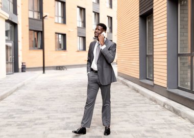 Takım elbiseli genç Afro-Amerikan erkek girişimci ofisin yakınında yürüyor, akıllı telefonla konuşuyor ve gülümsüyor, iş konuşması yapıyor, iş merkezi geçmişi, serbest alan