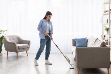 Tam vücut boy görüntüsü, günlük kıyafetlerle gülümseyen genç bir kadın ısıtılmış ahşap kaplama sıcak zemini mikrofiber ıslak bez kullanarak yıkıyor, ev ödevi temizliği yapıyor. Temizlik İşi Konsepti
