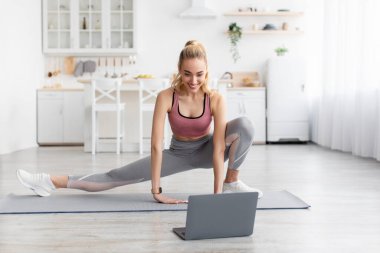Kafkasyalı güzel sarışın atletik kadın bacak egzersizleri yapıyor ve evde dizüstü bilgisayarla spor yapıyor. COVID-19 salgını, spor ve cihaz sırasında uzaktan ders.