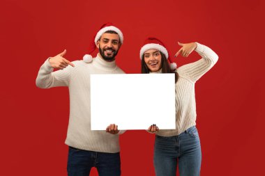 Neşeli Arap çift kırmızı Noel Baba şapkaları giyer ve kameraya gülümserken reklam için beyaz bir tahta tutarlar..