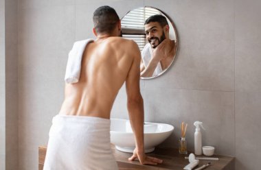 Neşeli bir Arap erkeği banyoda durmuş, sakalına dokunurken aynada kendine bakıyor. Bakımından sonra havluya sarılıp görünüşünden zevk alıyor..