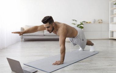 Coronavirus sırasında online spor. Genç ve zinde bir Arap, dizüstü bilgisayarın yanında egzersiz yapıyor, kalas pozisyonunda duruyor, tam boy. Orta Doğulu bir adam içeride video dersi veriyor.