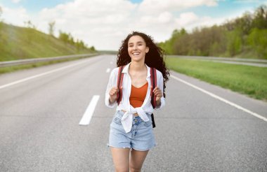 Yazlık giysiler içinde neşeli genç bir bayan kırsal alanda asfalt yolda yürüyor, dışarıda otostop çekiyor. Sırt çantalı, otostop ile seyahat eden güzel bir milenyum kadını.