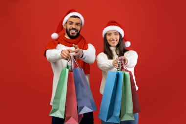 Noel Baba şapkası takan neşeli bir Arap çift ellerinde renkli alışveriş torbalarıyla kırmızı arka planda duruyor. Noel satışları ve indirimleri için alışveriş yaparak eğleniyorlar..