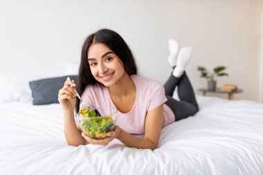 Sağlıklı beslenme, diyet ve detoks konsepti. Güzel Hintli kadın taze sebze salatası yiyor, yatakta uzanıyor. Güzel Asyalı bayan, içeride sağlıklı yemeğin tadını çıkarıyor.