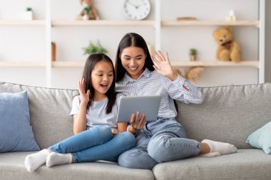 Modern uzaktan iletişim konsepti. Heyecanlı Asyalı anne ve kız dijital tablette video çağrısı yapıyorlar, kanepede oturuyorlar ve ekranda el sallıyorlar, internette sohbet ediyorlar.