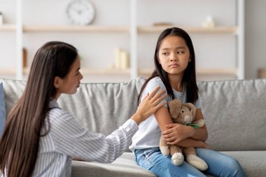 Aile içi çatışma kavramını çözmek. Genç Asyalı anne küçük kızı rahatlatıyor, onu teselli ediyor ve konuşmaya çalışıyor, evdeki kanepede oturuyor, boşluğu kopyalıyor.