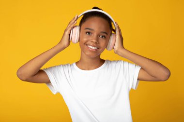 Beyaz tişörtlü, kablosuz kulaklıklı, gülümseyen, Afro-Amerikan kız müzik ve dans dinliyor, sarı arka planda izole edilmiş, kopyalama alanı. Olumlu yüz ifadesi, boş zaman ve rahatlama