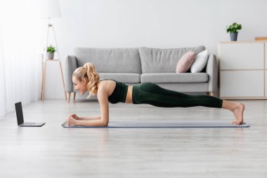 Üzgün Avrupalı güzel, bin yıllık sarışın bayan dizüstü bilgisayar başında video dersi alıyor, boş alan, profil. Çevrimiçi antrenör, spor egzersizleri uzaktan, oturma odasında ev içi spor salonu.