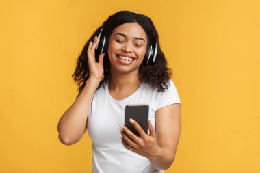 Modern teknolojiler. Kablosuz kulaklıkla müzik dinleyen Afrikalı Amerikalı kadın sarı arka planda akıllı telefondan şarkı arıyor. Siyahi kadın müzik listesinden hoşlanıyor