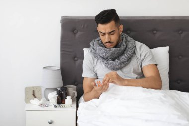 Genç bir Orta Doğulu adam kendini kötü hissederken ilaç alırken atkıya sarılmış bir şekilde yatakta oturuyor. Hastalığını gösteren dokular ve çeşitli ilaçlarla dolu..