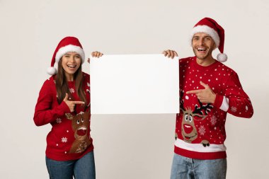 Renkli Noel süveteri giymiş iki gülen kişi, boş bir tabelayı işaret ederek yan yana duruyorlar. Bayram sezonunda şenlikli şapkalar giyer ve neşe saçarlar..