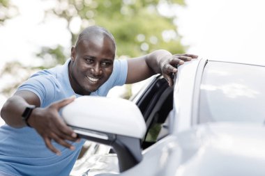 Araba perakendecisi, oto satın alma konsepti. Neşeli siyah adam günlük kıyafetleriyle dışarıda beyaz lüks arabanın yanında dikiliyor, yan aynaya bakıyor ve gülümsüyor, yeni bir araba seçiyor ve satın alıyor.