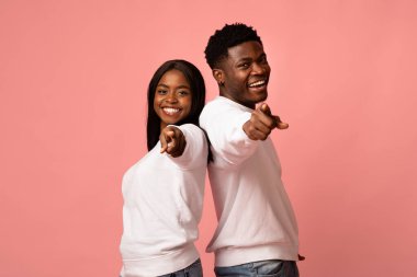 Neşeli bir Afro-Amerikan erkek ve kadın sırt sırta verip aynı kıyafetleri giyiyorlar. Yaygın bir şekilde gülümsüyorlar ve her ikisi de pembe arka planda işaret parmaklarıyla kamerayı işaret ediyorlar..