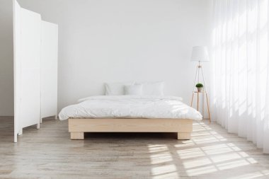Modern design, simple, modern home and domestic life. Real photo. Double bed on wooden floor in morning light from window with curtains, shield and lamp, on white wall background in bedroom interior