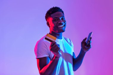 A cheerful young black man is holding a cellphone and a credit card while using a banking or shopping app. The vibrant neon light around him enhances the atmosphere of online transactions.