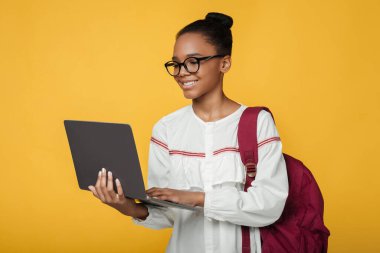 Sırt çantalı, sırt çantalı, dizüstü bilgisayarlı, sarı arka planda izole bir şekilde sınava hazırlanan zeki, güzel siyah kız öğrenci. Çevrimiçi ders sınıfı uzaklığı ve COVID-19 sırasında yeni normal