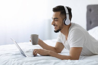 Bin yıllık Arap bir adam dizüstü bilgisayarını kullanıp kahvesini yudumlarken gülümseyerek yatakta dinleniyor. Kablosuz kulaklık takıyor ve boş zamanlarında internette sörf yapmaktan hoşlanıyor..