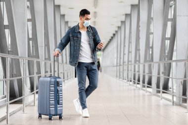 Havaalanında tıbbi maske takan, elinde bavuluyla terminalde dikilen, uçuşu beklerken cep telefonuna göz gezdiren, kopyalayan akıllı telefon kullanan genç bir adam.