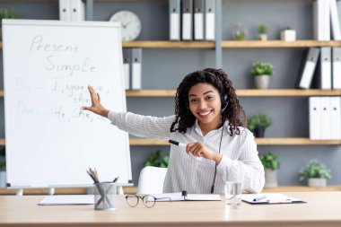 Neşeli genç bayan öğretmen tahtaya işaret ediyor, gramerini açıklıyor, ev ofisinden çevrimiçi İngilizce dersi veriyor. Kulaklıklı Afrikalı Amerikalı kadın webcam 'e bakıyor, içeride blog için çekim yapıyor.
