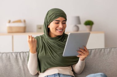 Genç bir Arap bayan, sıcak oturma odasındaki kanepede oturup başarısını neşeyle kutluyor. Bir tablet tutuyor ve heyecanla el kol hareketleri yapıyor. Mutluluğunu ve başarısını yansıtıyor..