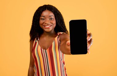 Güzel mobil alışveriş uygulaması. Portakal rengi stüdyo arka planında boş ekranlı akıllı telefon gösteren mutlu siyah kadın, tasarım için model. Kadın müşteri çevrimiçi bankacılık uygulaması öneriyor, seçici odak