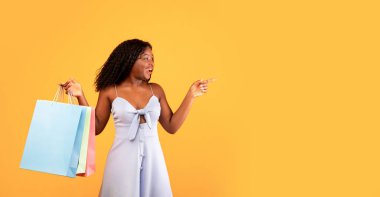 Parlak alışveriş torbaları olan heyecanlı genç siyahi kadın turuncu stüdyo arka planında büyük indirim ya da beklenmedik tanıtım öneriyor. Mevsimlik indirimler, teklifler, anlaşmalar