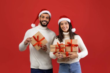 Neşeli bir Arap çift, ellerinde paketlenmiş Noel hediyeleri olan parlak kırmızı bir arka plana karşı birlikte duruyorlar. Noel Baba şapkası takıp kameraya gülümsüyorlar. Bayramların şenlik ruhunu paylaşıyorlar..