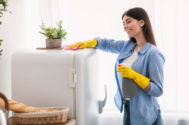 Temizlik ve Dezenfeksiyon Konsepti. Koruyucu lastik eldivenlerle gülümseyen bin yıllık kadın mutfakta duran beyaz buzdolabı yüzeyini siliyor. Neşeli bayan evini düzenli tutuyor, deterjan kullanıyor.