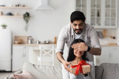Doğum günü sürprizi, yıldönümü, sevgililer günü ve evde tatil, covid-19. Neşeli, çekici genç Arap adam karısına gözlerini kapar ve oturma odasındaki koltukta kırmızı kurdeleyle hediye verir.