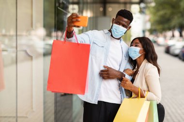 Karantina ve Salgın sırasında alışveriş. Tıbbi maske takan, alışveriş merkezinin yakınında selfie çeken, alışveriş torbası tutan ve cep telefonunun kamerasına gülümseyen mutlu Afro-Amerikan çiftin portresi.