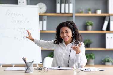 Çevrimiçi ders için modern teknoloji, eğitim, yeni normal, covid-19, uzak okul. Bin yıllık gülümseyen siyah kulaklıklı kadın öğretmen, tahtayı işaret ediyor, öğrenciyle konuşuyor, kapalı mekan kameralarına bakıyor.