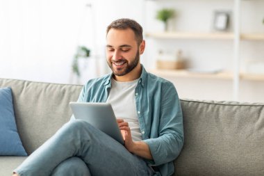 Neşeli milenyum adamı elinde tablet bilgisayarla kanepede oturuyor, çalışıyor ya da evde internetten çalışıyor. Web konferansına katılan mutlu beyaz erkek, webinar veya video konferansına katılıyor