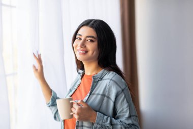 Genç bir Arap kadın pencerenin kenarında bir fincan kahve tutarken gülümsüyor, evde rahatlatıcı bir sabahın tadını çıkarıyor. Doğal ışık perdelerin arasından süzülerek sıcak bir atmosfer yaratır..