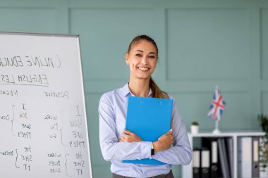 Neşeli bir öğretmen sanal bir İngilizce dersine liderlik ediyor, video aracılığıyla öğrencilerle iletişim kurarken gülümsüyor. Elinde bir pano var ve eğitim içerikli bir beyaz tahta tarafından yerleştirilmiş..