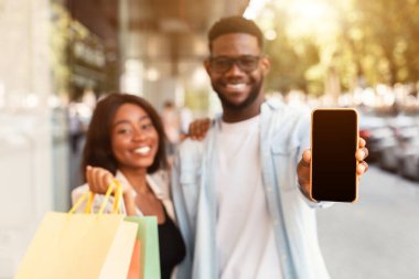 Çevrimiçi Mağaza Uygulaması. Mutlu siyah çift kameraya yakın boş ekranlı akıllı telefonları gösteriyor ve ellerinde çantalar, dışarıda duran neşeli insanlar, Mock Up, bedava fotokopi alanı.