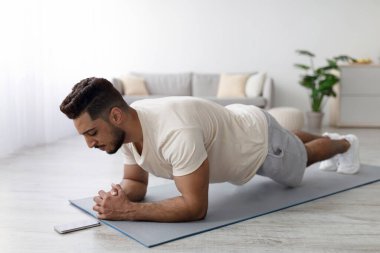 Ev içi güç egzersizi. Genç Arap adam evde dirsek kalası yapıyor, cep telefonundaki video dersini takip ediyor. Yakışıklı Orta Doğulu adam kapalı alanda eğitim görüyor.