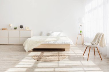 Beyaz yatak odasında sabah ışığı. Yastıklı yatak, ahşap zeminde halı, perdeli pencere, mobilya, sandalye ve dekor elementleri yatak odası arka planında, boş alan, kapalı alan