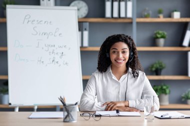 Çevrimiçi öğretmen, uzaktan çalışma, evde modern eğitim, covid-19 salgınında sosyal mesafe. Milenyum Afro-Amerikan kadın kulaklıklı kara tahta arka planda web kamerasına bakıyor.