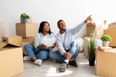 Heyecanlı Afro-Amerikan çifti dijital tabletleri ile yeni evlerine ev eşyaları almak için internette geziniyorlar. Yeni evliler taşınma gününde karton kutuların arasında oturmuş iç mekan hayal ediyorlar.