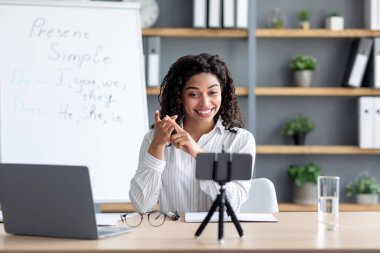 Uzaktan eğitim, sanal toplantı, webcam 'e video çağrısı, evde öğrenim ya da öğretmenlik. Pozitif genç siyahi bayan öğretmen, arka planda, jest, açıklamalar kurallar, kapalı alanda telefona bak.