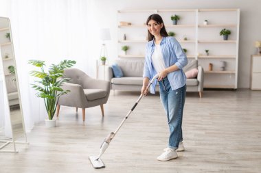 Ev ödevi temizleme rutini. Neşeli, sıradan bir kadın, ahşap döşemeleri yıkıyor, su püskürtme pedi kullanıyor, gülümsüyor ve kameraya bakıyor. Bulanık Arkaplan, Serbest Kopya Alanı, Modern İçsel