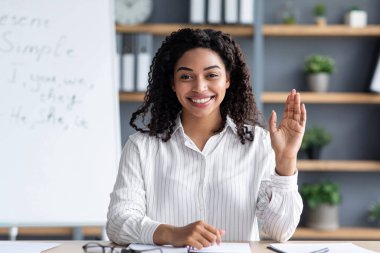 Covid-19 salgını sırasında çevrimiçi dil okulunda. Bloglar için sosyal mesafe ve teknoloji. Kara tahtanın arka planında mutlu siyah bir kadın el sallayan ve ateş eden web kameralarına bakıyor.