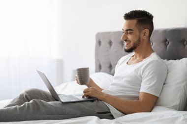 Neşeli genç bir Orta Doğulu adam bir dizüstü bilgisayar ve bir fincan kahveyle yatakta dinleniyor. Sabah haberlerini okurken gülümsüyor, rahat yatak odasının keyfini çıkarıyor ve güne huzurlu bir başlangıç yapıyor..