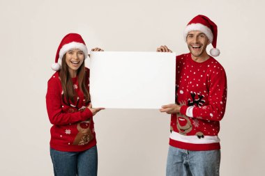 Neşeli bir çift Noel süveteri ve şapkaları giyer birlikte durur, gülümser ve boş bir tabela tutarlar. Arka plan, neşeli ifadelerini ve tatil ruhlarını vurgulamak için açık..