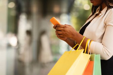 Tüketim ve Teknoloji. Tanımlanamayan siyah bir kadının akıllı telefon kullanması, alışveriş torbalarını tutması, dışarıda durması, kıyafet seçip sipariş etmesi, hediyeler alması, fotokopi alanı, bulanık arka plan.