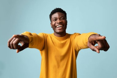 Mavi stüdyo arka planında poz veren, neşeli gülümseyen ve kameraya el kol hareketi yapan Afro-Amerikan genç adam işaret parmağı gösteren, flört eden, kopyalayan