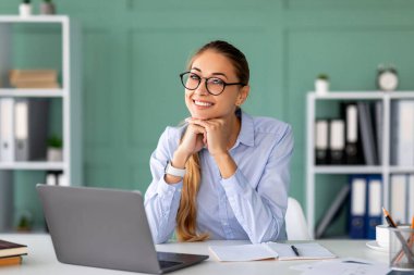 Genç bayan CEO, modern bir ofiste çalışma odasında oturuyor, dizüstü bilgisayarı üzerinde çalışırken mutlu görünüyor. Parlak iç mekan, yoğun gününün neşeli atmosferini arttırır..