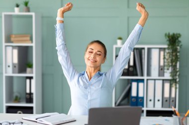Modern bir ofiste masa başında oturan kadın bir ofis çalışanı stresten kurtulmak için kollarını kapalı bir şekilde uzatıyor. O, yoğun gününde dizüstü bilgisayarı üzerinde çalışırken rahat görünüyor..