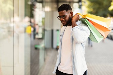 Gözlüklü yakışıklı zencinin portresi moda kıyafetlerine bakıyor ya da alışveriş merkezinin vitrininde yeni ürünlere bakıyor, alışveriş merkezinin yakınında çantalarını tutuyor. Hediyeler, Satış ve İndirim, Perakende, Satın Alma Konsepti